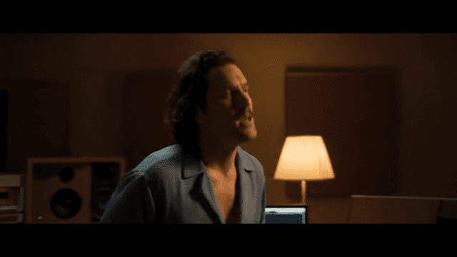 A male singer in his 30s with medium-length curly hair, a thin mustache, and a soulful gaze stands in a warm 1980s-style recording studio. He wears a loose open-collar shirt and softly sways while singing the lyric: "We are the prompts". The scene has a soft VHS grain filter, warm amber lighting, and is filmed in a vintage 4:3 aspect ratio. The cinematography mimics a music charity anthem from the 1980s, with a slow push-in toward the singer's face, highlighting his emotional expression.