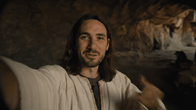 A cinematic handheld selfie-style video shot, showing a young man in ancient Middle Eastern robes with shoulder-length dark hair and a short beard, holding the camera like a selfie cam. He's inside a dimly lit stone den with rugged cave walls. Behind him, several large lions slowly pace or rest, casting shadows in the flickering torchlight. The man speaks directly into the camera he holds, slightly amused expression as he whispers.
He says: Alright, welcome to my crib. That one is asleep, he gets grouchy in the mornings.
He looks happy as he whispers, as if he knows a secret. He pans the camera to show the lions in the background behind him.
The light pours from the cave entrance above that illuminates the dark cave chamber with the lions below.
Time of Day: day
Lens: ultra-wide selfie lens, shallow depth of field
Film Stock: vintage 35mm documentary style, selfie camera view
Audio: (implied) ambient lion growls
Background: Lions sleeping behind the young man.